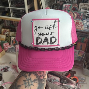 Pode incluir: Boné de caminhoneiro rosa e branco com o texto "go ask your DAD" em preto. O boné tem uma aba e malha rosa, com um detalhe de corrente preta na frente.