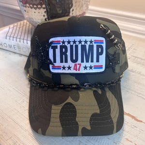 May include: A camouflage trucker hat with a black chain trim and a patch that says "TRUMP 47" with stars and red, white, and blue stripes.