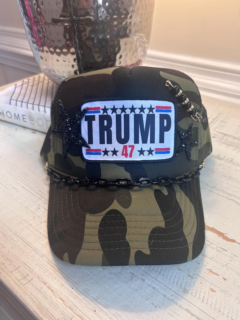 May include: A camouflage trucker hat with a black chain trim and a patch that says "TRUMP 47" with stars and red, white, and blue stripes.