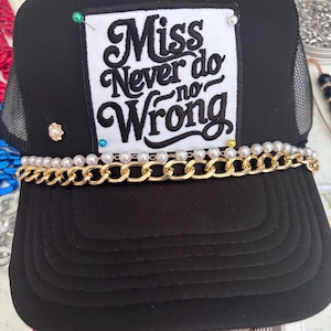 Pode incluir: Boné de caminhoneiro preto com parte traseira em malha e aba plana. A frente apresenta um patch branco com o texto "Miss Never do no Wrong" em escrita preta. Uma corrente dourada e pérolas adornam a aba, com alfinetes decorativos.