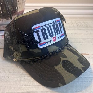 May include: A camouflage trucker hat with a black chain and a red, white, and blue patch that says "TRUMP 47***".