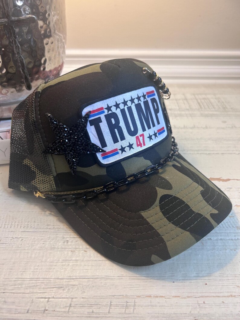 May include: A camouflage trucker hat with a black chain and a red, white, and blue patch that says "TRUMP 47***".