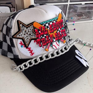 May include: A baseball-themed trucker hat with a black brim and a white and black checkered pattern on the sides. The hat features a patch with the text "ALL ABOUT THAT BASE" and a star, baseball, and bats.