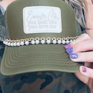Pode incluir: Boné de caminhoneiro verde com um remendo branco que diz "Cause Me, You Look Like You Love Me". O boné tem uma corrente dourada e uma borda de pérolas.