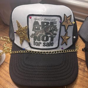 Pode incluir: Boné de caminhoneiro preto e branco com parte traseira em malha e aba curva. O painel frontal apresenta um patch com um tigre e o texto "GOD'S CHILDREN ARE NOT FOR SALE". Estrelas douradas e detalhes de corrente completam o design.