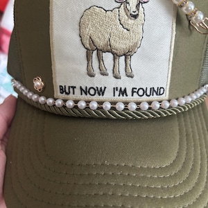Pode incluir: Boné de caminhoneiro verde-oliva com um patch creme bordado com uma ovelha e o texto "BUT NOW I'M FOUND". O chapéu é adornado com pérolas e um alfinete decorativo.