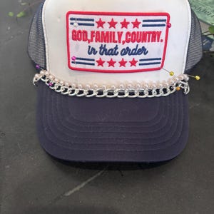 Pode incluir: Boné de caminhoneiro azul marinho e branco com o texto "GOD, FAMILY, COUNTRY. in that order" em um patch vermelho e azul. O boné apresenta uma corrente prateada e enfeites de pérolas ao longo da aba.