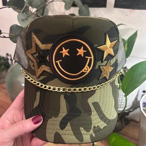 Pode incluir: Um boné de caminhoneiro camuflado com uma corrente dourada, um remendo de rosto sorridente preto com estrelas douradas e dois remendos de estrelas douradas.