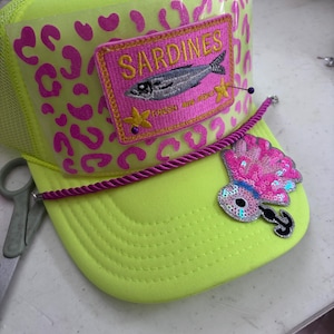 Pode incluir: Boné de caminhoneiro amarelo neon com estampa de leopardo rosa. O boné apresenta um patch com a palavra "Sardinhas" e uma ilustração de peixe. Um cordão rosa e roxo adorna a aba, com um enfeite de lantejoulas.