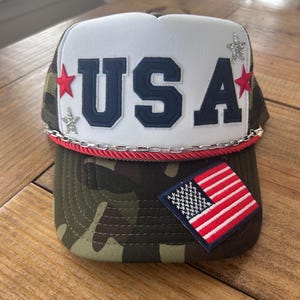 Pode incluir: Um chapéu de caminhoneiro branco e camuflado com um remendo de bandeira americana vermelho, branco e azul e a palavra "USA" em letras azuis. O chapéu tem uma corrente prateada e uma guarnição de corda vermelha.