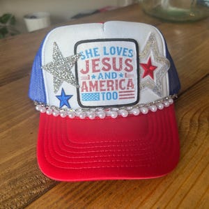 Pode incluir: Um chapéu de caminhoneiro branco, azul e vermelho com um remendo que diz "She Loves Jesus and America Too". O chapéu tem uma estrela prateada, uma estrela vermelha e uma estrela azul. O chapéu também tem uma fileira de pérolas brancas ao redor da borda.