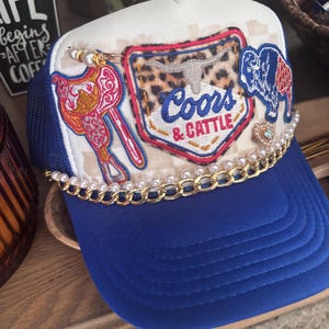 Pode incluir: Boné de caminhoneiro azul e branco com aba azul. O boné apresenta patches bordados com uma sela, um longhorn e um bisonte. O texto "Coors & Cattle" é bordado em um patch. O boné é decorado com pérolas e uma corrente dourada.