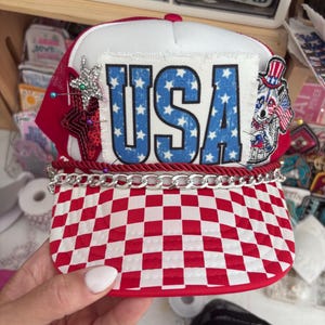Pode incluir: Um boné de caminhoneiro vermelho, branco e azul com a palavra "USA" em letras azuis com estrelas brancas. O boné tem uma viseira xadrez vermelha e branca, uma corrente prateada e um patch de cachorro patriótico.