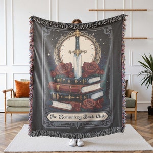 May include: A dark gray woven throw blanket featuring a fantasy design. The design includes a sword, roses, and a stack of books. The text "The Romantasy Book Club" is at the bottom of the design. The blanket has a fringed edge.