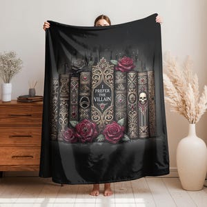 May include: Black throw blanket featuring a design of antique books with gold accents, red roses, and a skull. The text "I Prefer The Villain" is displayed on one of the books. The blanket is held up in front of a person.