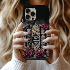 May include: Black phone case featuring a design of antique books, red roses, and the text "I Prefer the Villain." The case has a matte finish and a detailed, gothic aesthetic. The roses are a deep red color.