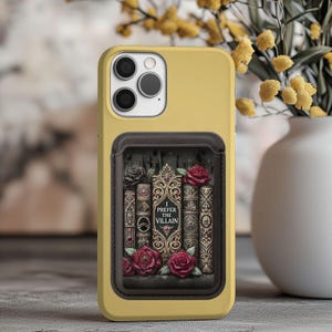 Puede incluir: Una funda de smartphone color amarillo mostaza con un tarjetero rectangular marrón oscuro en la parte posterior. El tarjetero muestra una ilustración de libros antiguos con detalles dorados, rosas rojas y negras, y el texto I PREFER THE VILLAIN.