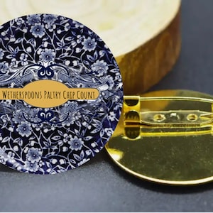 Puede incluir: Un pin redondo con un diseño floral azul y blanco y el texto "Wetherspoons Paltry Chip Count" en un óvalo dorado. El pin tiene un respaldo dorado con un imperdible.