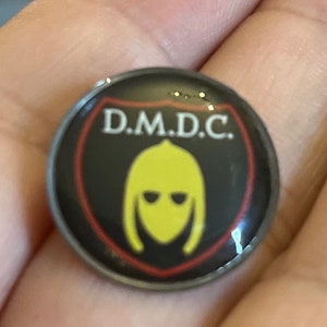 Puede incluir: Un pin circular negro y rojo con un casco amarillo y el texto "D.M.D.C."