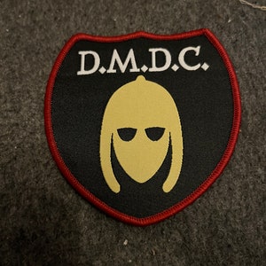 Op de afbeelding: Zwarte en rode geborduurde patch met de tekst "D.M.D.C." en een geel gestileerd gezicht met een zonnebril.