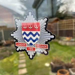 Puede incluir: Una pegatina en blanco y negro con un escudo rojo y azul. El escudo tiene tres coronas y líneas onduladas. La pegatina dice "London Fire Brigade".