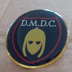 Puede incluir: Un pin redondo y negro con borde dorado. El pin presenta un diseño de escudo con las letras "D.M.D.C." en blanco en la parte superior. Dentro del escudo hay una figura amarilla con casco y ranuras oscuras para los ojos. El escudo está delineado en rojo.