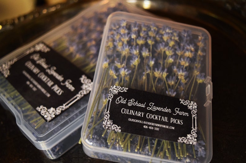 May include: Two clear plastic containers filled with dried lavender sprigs. The containers have black labels with white text that reads "Old School Lavender Farm Culinary Cocktail Picks" and includes the farm's email address and phone number.