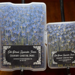 May include: Two clear plastic containers filled with dried lavender flower sprigs. The containers are labeled "Old School Lavender Farm Culinary Cocktail Picks". The larger container holds sprigs that are approximately 6 inches long, while the smaller container holds sprigs that are approximately 4 inches long.