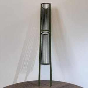 Puede incluir: Un soporte decorativo alto de metal verde oliva para plantas con un diseño de listones verticales. El soporte presenta un marco rectangular con una serie de barras verticales y detalles curvos. El soporte mide aproximadamente 152 cm de altura.