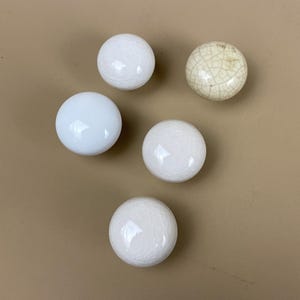 May include: Five white ceramic cabinet knobs. Four have a crackle glaze finish, and one is smooth. The knobs are round and vary in colour, with a creamy off-white and a bright white.