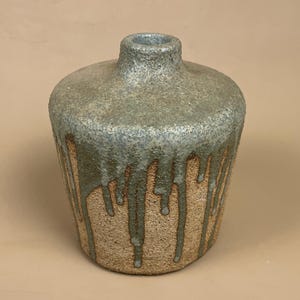 May include: A handmade ceramic vase with a textured, speckled finish. The top section is a muted blue-green, with a dripped glaze effect in a similar hue cascading down over a tan base. The vase has a rounded body and a narrow neck.