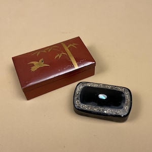 May include: Two decorative boxes. One is rectangular, red, and decorated with a gold bird and bamboo design. The other is black with a silver border and a small, oval, iridescent accent in the centre. Both boxes are closed.
