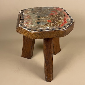 May include: A small, antique wooden stool with a square seat. The seat is upholstered with a patterned fabric in shades of gold, red, and brown, and is secured with metal studs. The stool has three sturdy, dark brown legs.