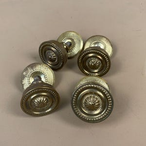 May include: A set of antique brass door knobs. Each knob has a circular design with a central floral pattern and a decorative outer ring. The knobs have a warm, aged patina, suggesting vintage charm.
