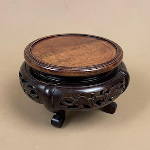 May include: A dark brown wooden display stand with a round top and carved floral design. The stand has three short legs and a circular top, ideal for displaying small decorative items. The wood has a rich, polished finish.