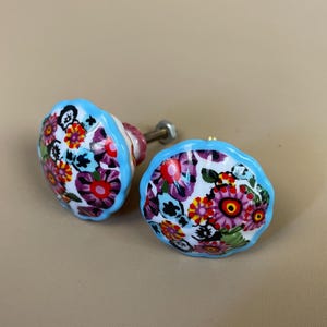 May include: Two floral cabinet knobs. Each knob features a white background with a colourful floral pattern in shades of red, purple, yellow, and green. The knobs have a light blue scalloped edge and a metal screw.
