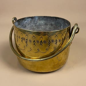 May include: A vintage brass bucket with a handle. The bucket has a round shape with a decorative floral pattern around the top. The brass has a warm, golden colour.