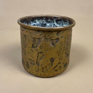 May include: A small, cylindrical brass container with etched designs. The container has a slightly tarnished appearance, with dark accents in the etched areas. The interior shows a greenish-blue patina. The exterior features etched figures and patterns.
