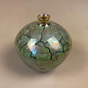 May include: A decorative, round vase with a cracked, iridescent green glaze. The vase has a gold-coloured rim and a unique, textured surface. The overall design is reminiscent of a mosaic or cracked earth.