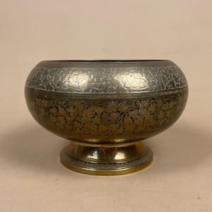 May include: A round metal bowl with a pedestal base, featuring a two-tone finish. The upper section has a silver-toned surface with intricate floral engravings, while the lower section displays a dark, tarnished gold-toned floral pattern.