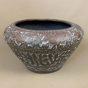 May include: A copper-toned, round bowl with detailed etched patterns. The bowl has a dark interior and a wide rim. The exterior is decorated with animal and foliage designs, creating a textured surface. The bowl is likely made of metal.
