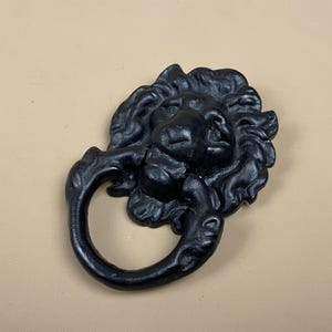 May include: A black, cast iron lion head door knocker. The lion's head has detailed features, including a mane and a ring-shaped handle. The door knocker is mounted on a beige surface.