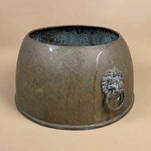 May include: An antique bronze planter with a weathered patina. The planter has a rounded shape and features a decorative lion's head with a ring handle on one side. The interior shows a greenish hue.