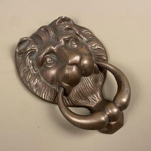 May include: Bronze lion head door knocker with a ring handle. The lion's face is detailed with fur and facial features. The door knocker is mounted on a light beige surface.