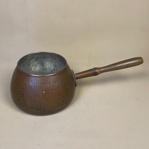 May include: A hammered copper saucepan with a wooden handle. The saucepan has a rounded bottom and a wide, shallow body.