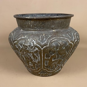 May include: Antique bronze-coloured metal pot with detailed relief carvings. The pot features a wide, rounded body and a slightly flared rim. The relief depicts figures and floral patterns, adding a decorative element to the vessel.