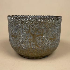 May include: An ornate metal bowl with etched patterns. The bowl has a silver and gold finish, with detailed designs of figures and floral elements. The rim is scalloped, enhancing its decorative appeal. The bowl is likely made of brass or a similar metal.