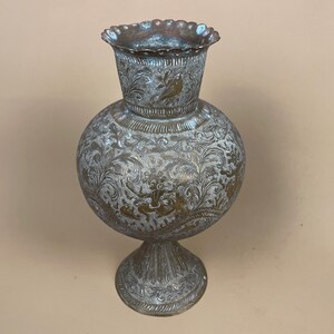 May include: An antique metal vase with detailed floral and bird engravings. The vase features a rounded body, a flared rim, and a pedestal base. The metal has a tarnished, aged look, with hints of gold and blue-green patina.
