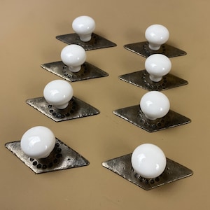 May include: Eight white ceramic cabinet knobs with a round shape, mounted on diamond-shaped, dark metal backplates. The backplates have a hammered texture and a decorative border of small, evenly spaced rivets. The knobs are arranged in a grid pattern.