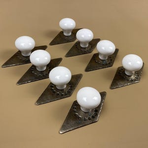 May include: Nine antique white ceramic knobs on diamond-shaped, aged metal backplates. The backplates have a hammered texture and are arranged diagonally. The knobs are round and smooth, creating a contrast with the textured metal.